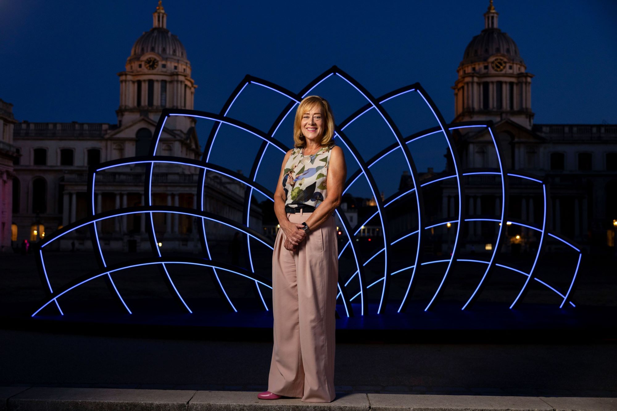 Vice-Chancellor Professor Jane Harrington wearing pink trousers and flower top smiles as she is stood on a step in front of an illuminated blues sculpture. Behind her you can see the two domes of the Old Royal Naval College lit up in the dark