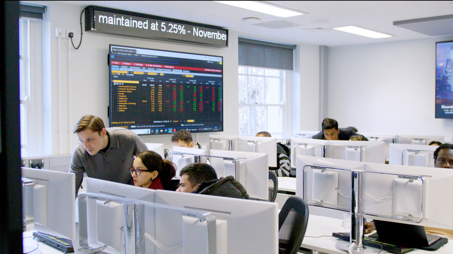 Tutor teaching two students who are staring to a desktop how to use the bloomberg terminals