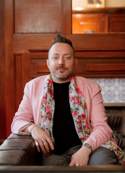 A person is sat on an armchair in a pink jacket, a pink floral scarf in front of a  wooden background