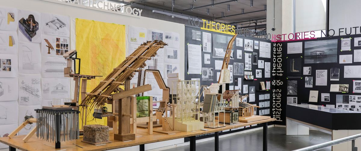 End of Year Exhibition showing the technology and thesis exhibition. The image features numerous detailed models and design drawings displayed on a long table. The walls are covered with various posters, blueprints, and imagery related to architecture.