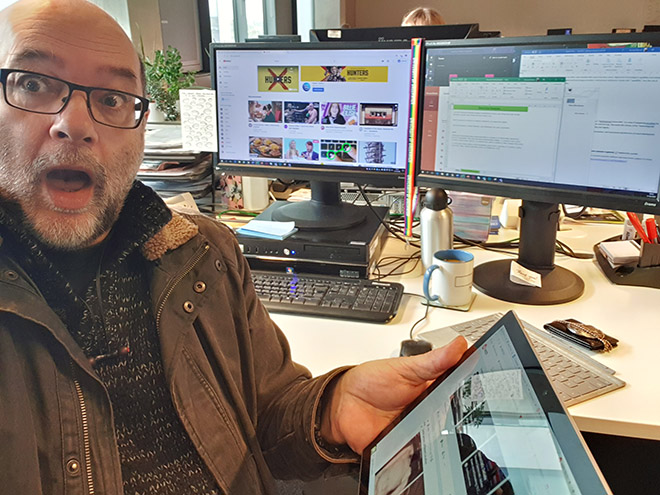 Mike is surrounded by tech, he is holding a tablet and has two computer screens behind him. He looks surprised. Mike is a white man wearing a brown coat.