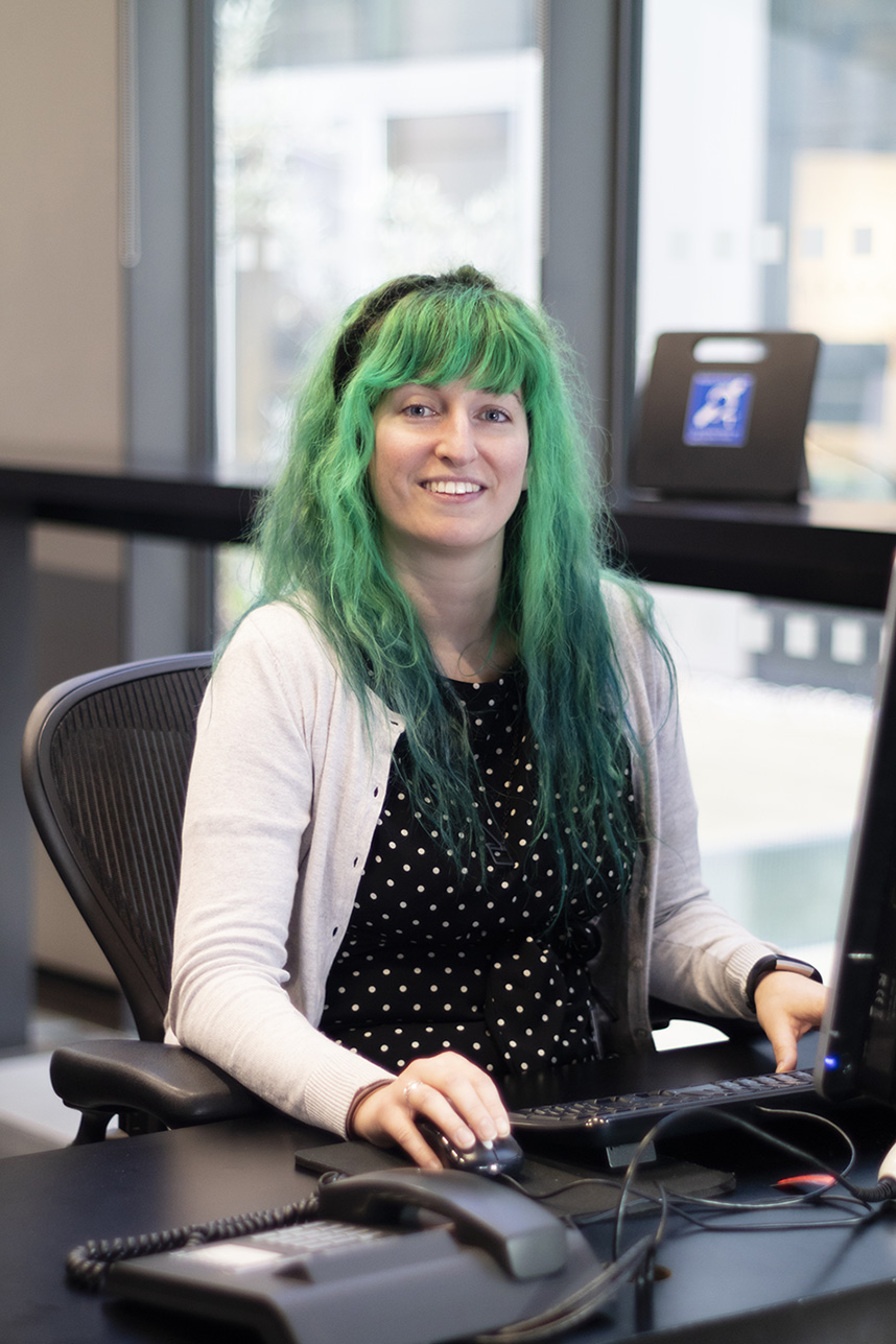 Maira sits at our library support desk and smiles at the camera. She is a white woman with bright green hair and wears a black top with cream jumper.