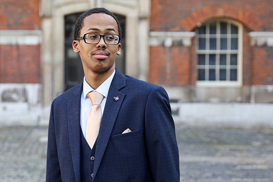 Abdirahman is in a blue suit and is stood in front of a building