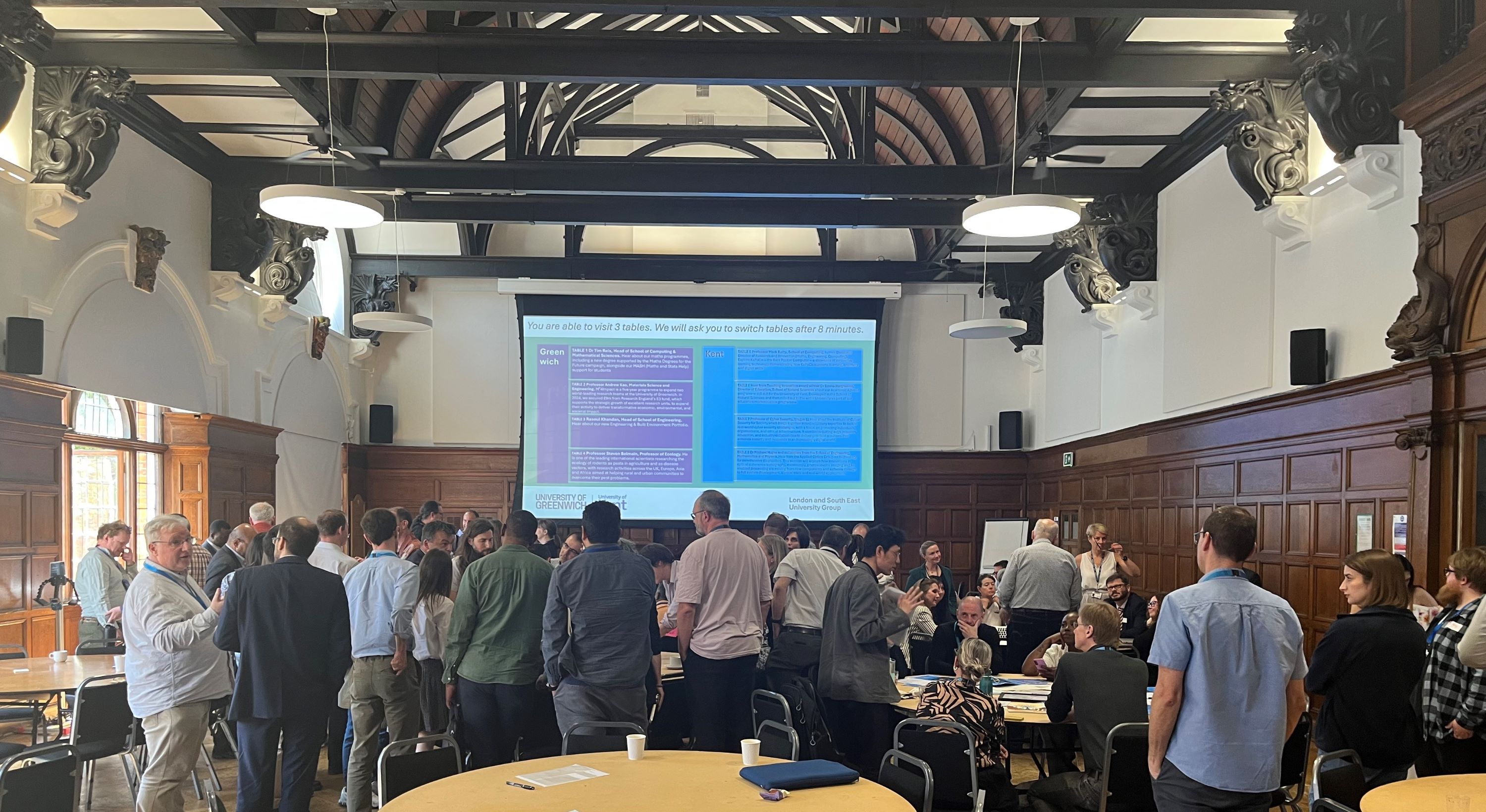  staff stood together talking in a large room, there is a presentation on the screen and staff look happy as they are chatting