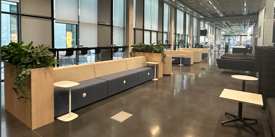 Stockwell street ground floor The ground floor of Stockwell Street Library, showing an open-plan seating area with upholstered bench seating, armchairs, small tables and integrated wooden planters filled with greenery. Floor-to-ceiling windows line one side of the space, and polished dark flooring extends through the seating area towards the library entrance.