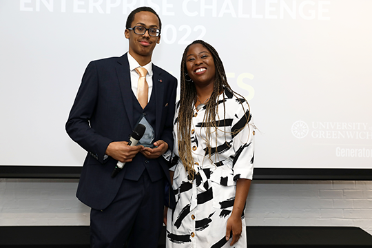 Abdirahman is in a blue suit holding a microphone and a glass award stood next to a woman wearing a white dress with black stripes