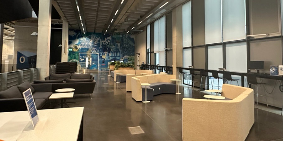 The ground floor of Stockwell Street Library, showing an open seating area with curved upholstered sofas, armchairs and small tables arranged across a polished floor. A row of study desks and computers runs alongside floor-to-ceiling windows, while a large blue mural covers the far wall of the space. Integrated planters with greenery are positioned between seating areas throughout the open-plan layout.