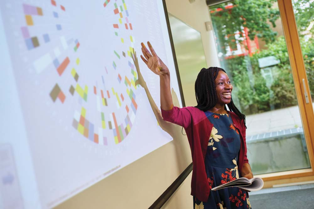 A black female lecturer stands in front of a projection on a whiteboard and smiles towards the room.