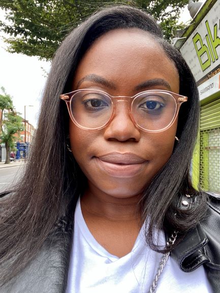 A person with glasses taking a selfie walking down the street. There are trees behind them.