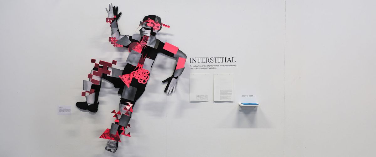 A photograph of student work on display in the degree show. A black silhouette of a human figure in a marching pose at near life-size cut out of card, mounted at head height on a plain white wall. Parts of the body showing skin are cut out of card and mounted on top of the black silhouette. On top of the body parts are mounted small interconnected geometric shapes in purple and bright pink. To the right on the wall is hung A4 white sheets of paper printed with text explaining the project. On a small shelf at stomach height, there is a pile of A6 blue cards labelled ‘yours to keep’.