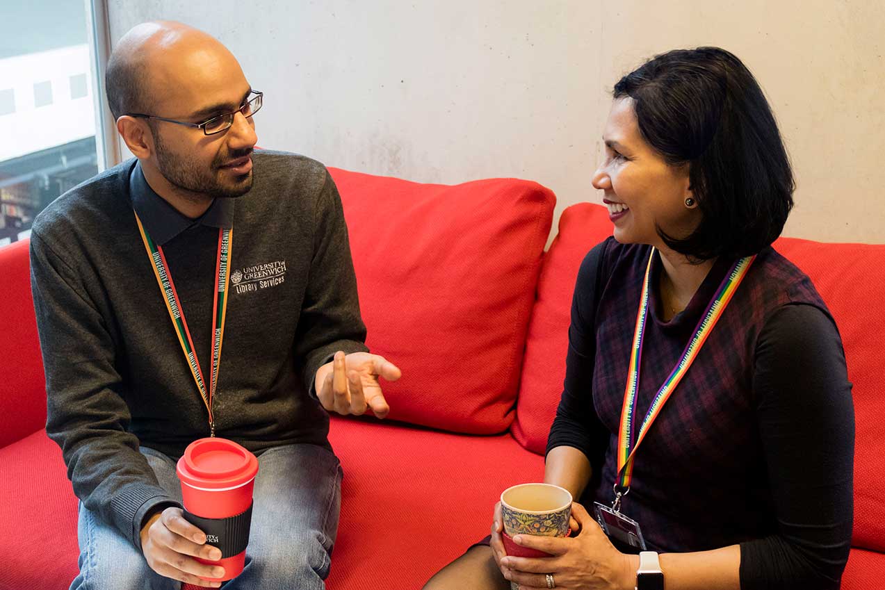 Vik and Sharon sit on a red sofa having a friendly conversation. Vik is an a british indian and wears a library hoodie, while Sharon is British South-Asian and wears a black top. They both hold cups of coffee.