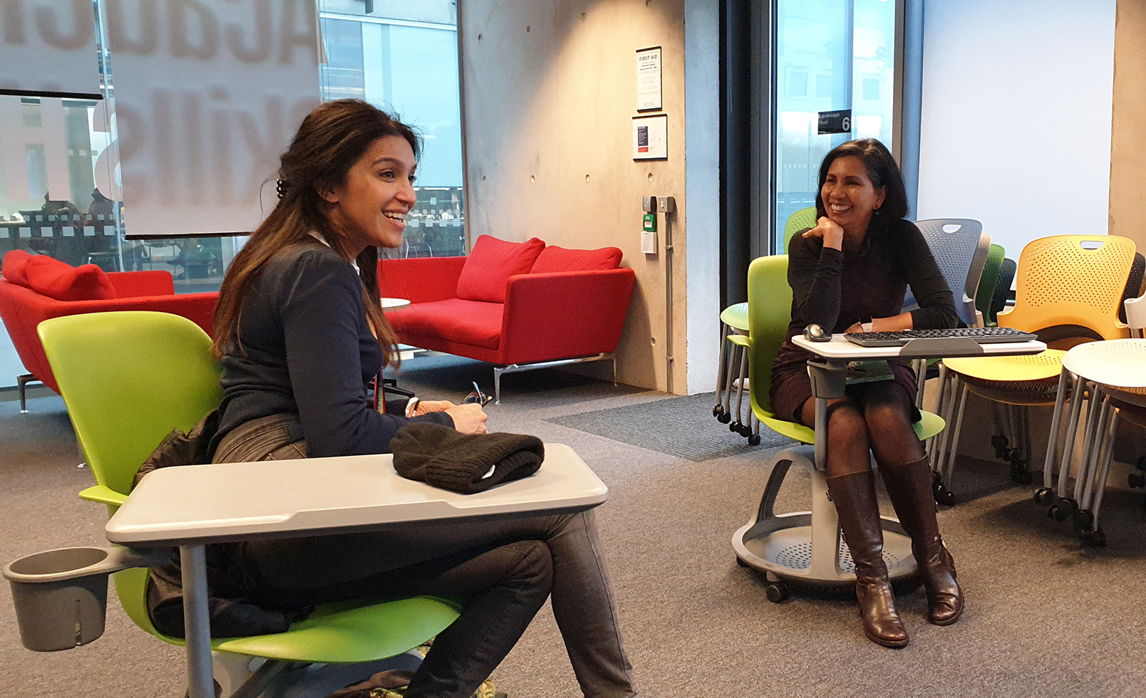 Two friends sit smiling in lecture chairs. They are south-Asian women wearing smart workwear.