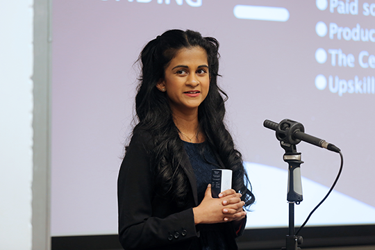 Shelini is stood holding a note card in front of a microphone. A dark screen is behind her