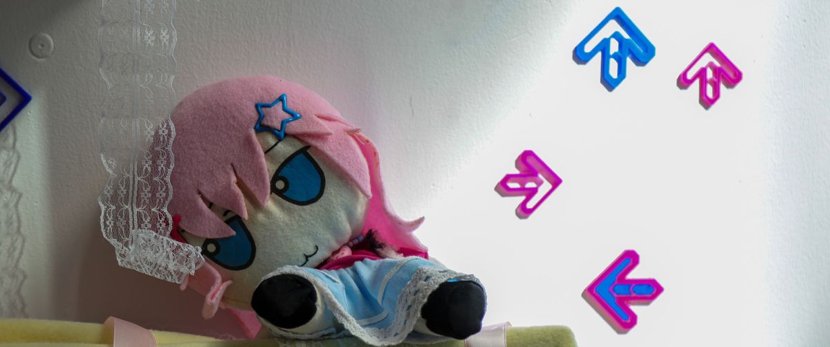 A photograph of student work on display in the degree show. In front of a plain white wall sits a plush Fumo doll slumped to the left in a shadow. The doll has pink hair made of felt with a blue star-shaped hair pin, large blue eyes, a white face, a blue skirt with white embroidered edging and black feet. A piece of white embroidered edging, like that on the skirt, hangs down from above in front of the doll's head. Slumped towards it, the doll appears to stare at it. To the right in brighter light are four small, short arrows laser-cut from pink and blue acrylic with two pointing up, one pointing right and the other pointing left. 