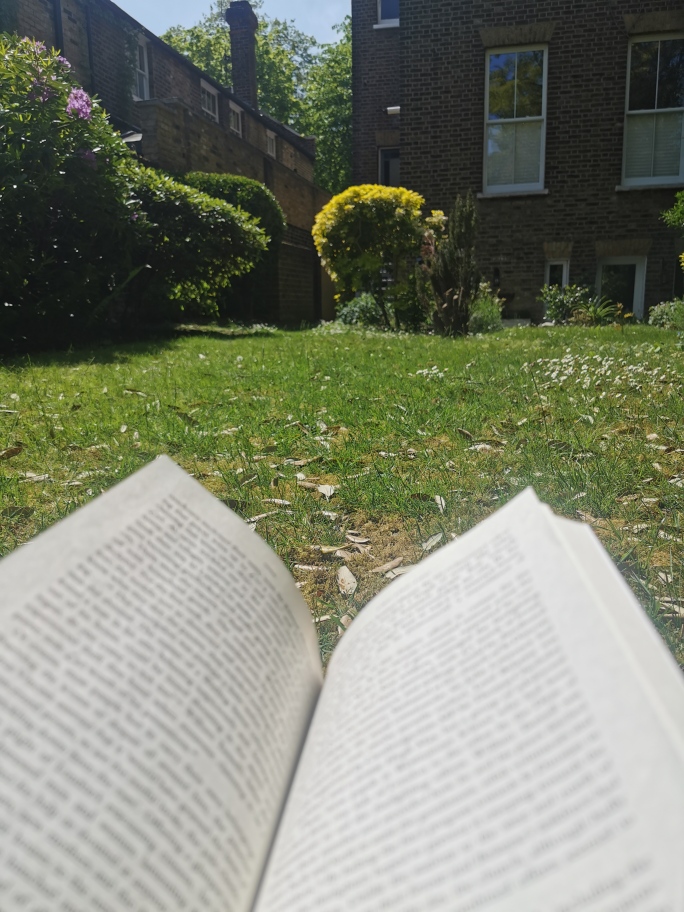 An open book in a garden, grass, bushes and a house in the background.