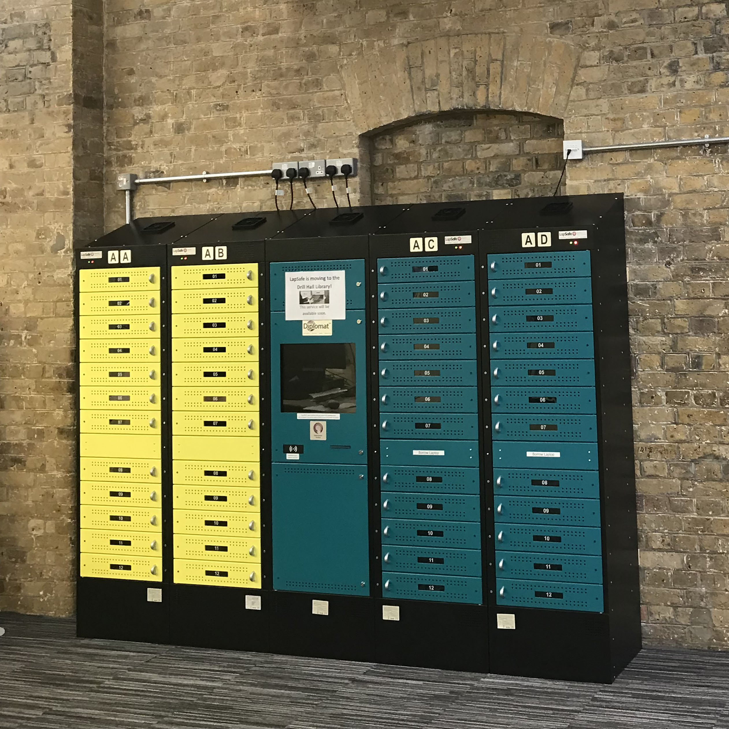 Lapsafe Locker in Drill Hall Library. The lockers are yellow and green with a black surround. It stands tall against an exposed brick wall.