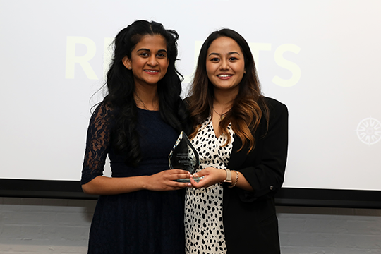 Shelini is stood in a black dress next to a woman in a black and white polka dot dress and black blazer. They are holding a glass award