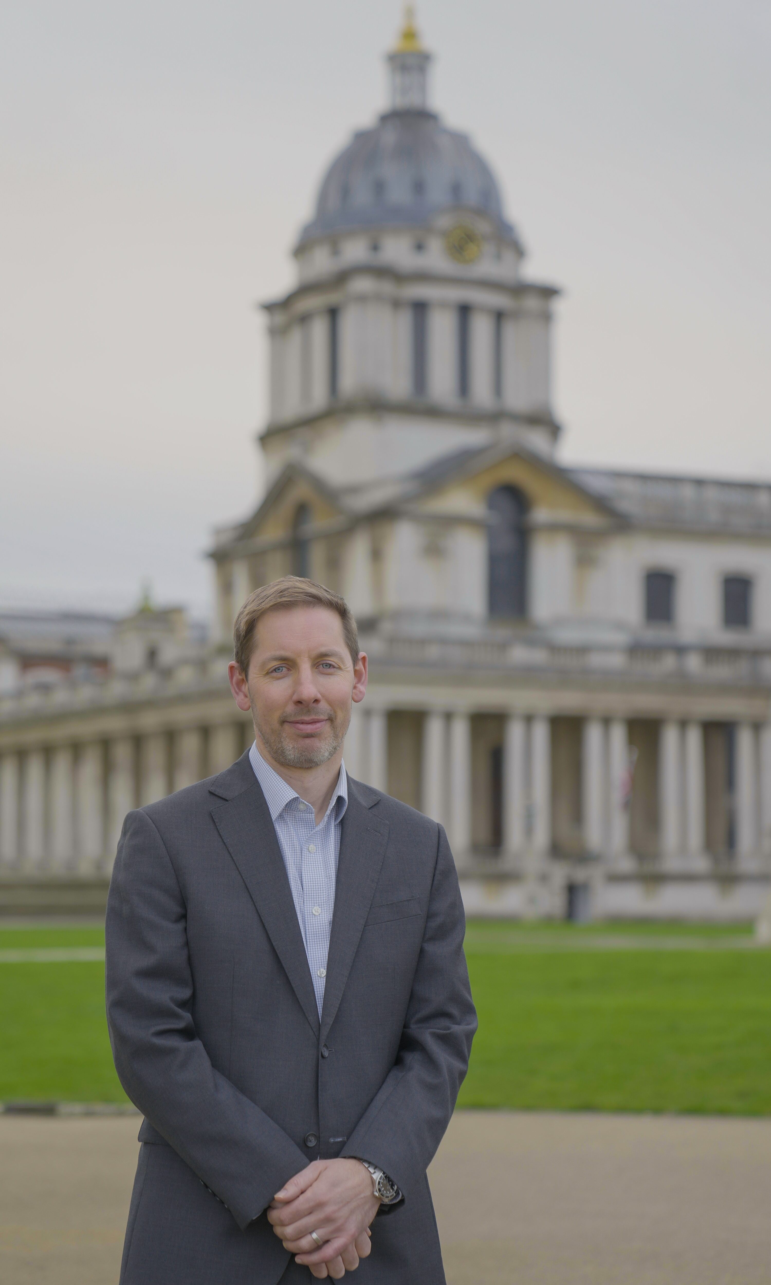 A person wearing a dark business suit and light shirt stands outdoors with hands clasped in front. In the background is the historic domed building of the Old Royal Naval College in Greenwich, London, with columns, a clock face, and landscaped lawns visible. The building is softly blurred, drawing focus to the person in the foreground.