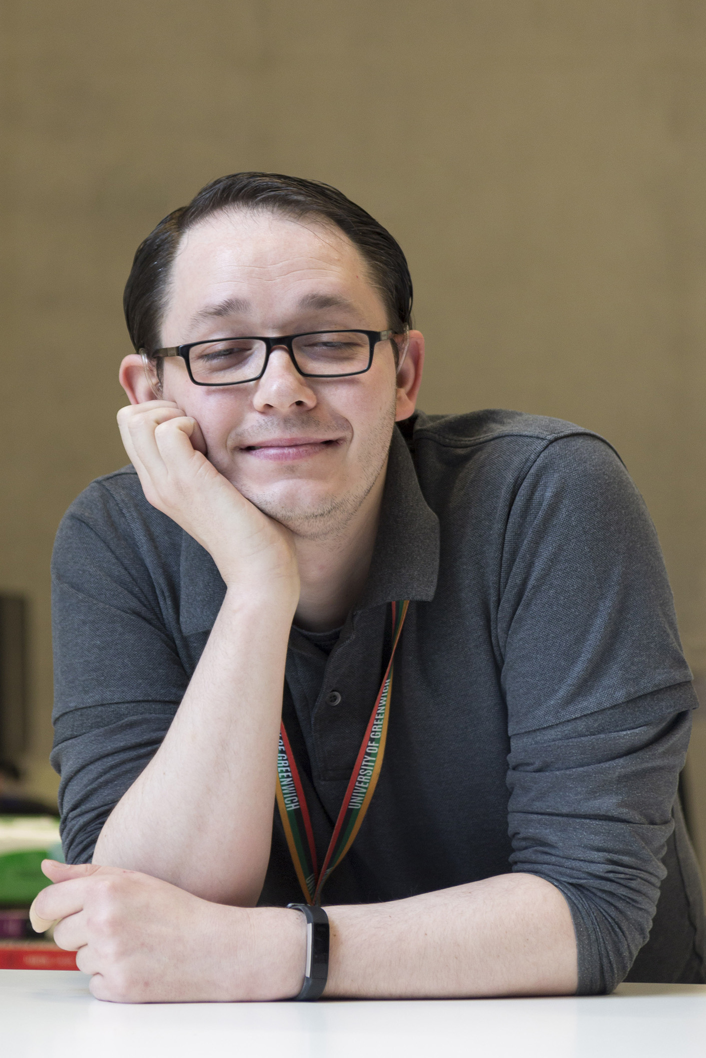 James leans one elbow on the desk and rests his head in that hand. He smiles at the camera. James is a white man wearing a grey top.