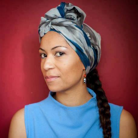 person in a light blue top and patterned headwrap with a long braid, posed against a red background