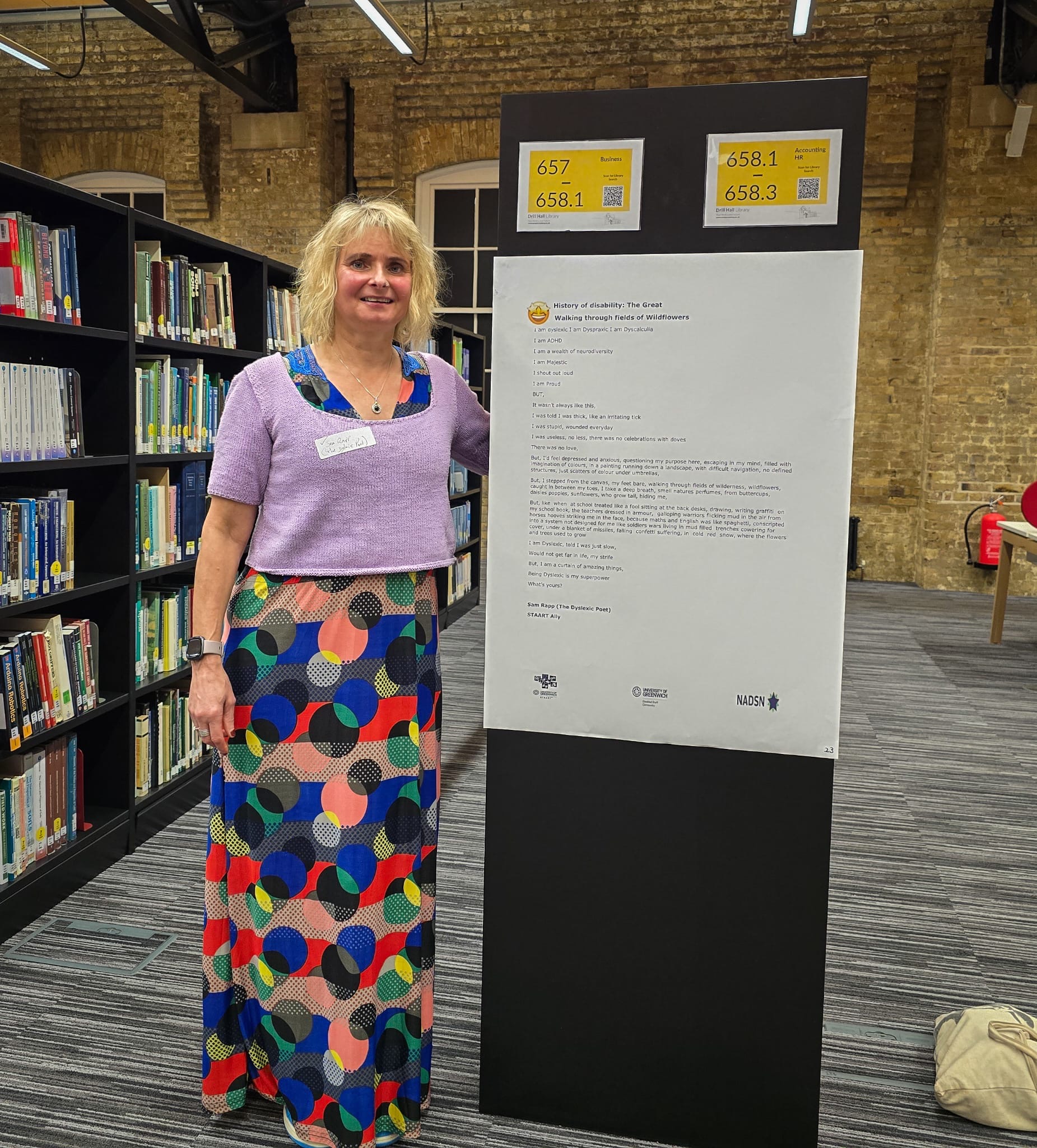 Blonde haired female dressed in a purple top and printed skirt stands next to an sketchboard in a library hall