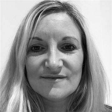Black-and-white head-and-shoulders portrait of a person with long, light-coloured hair, facing the camera against a plain indoor background. The image is closely cropped around the face and shoulders, with even lighting and a neutral expression.