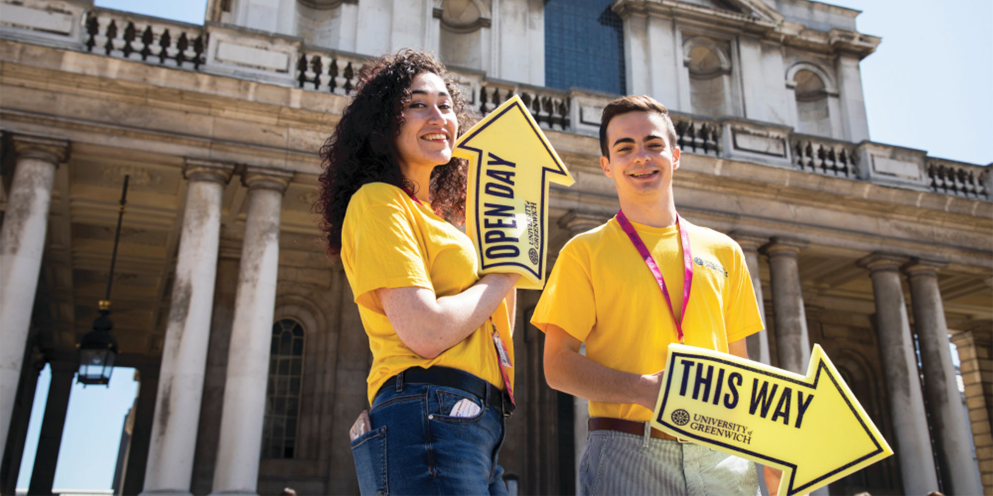 Two student ambassadors standing holding yellow arrow signs in front of the Old Royal Naval College