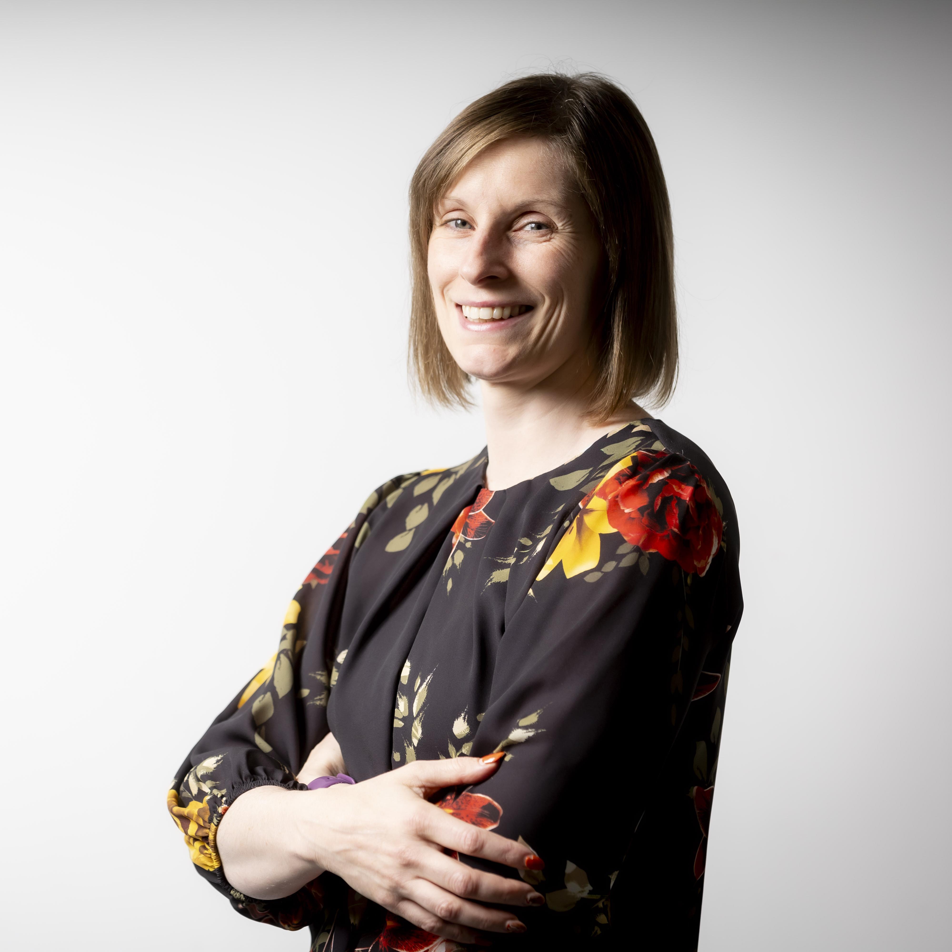 Studio portrait of a person standing against a light grey background, shown from the waist up with arms folded. The person has shoulder-length hair and is wearing a dark long-sleeved top with a bold floral pattern in red, yellow and green. The image is brightly lit with the subject positioned slightly to the right of the frame.