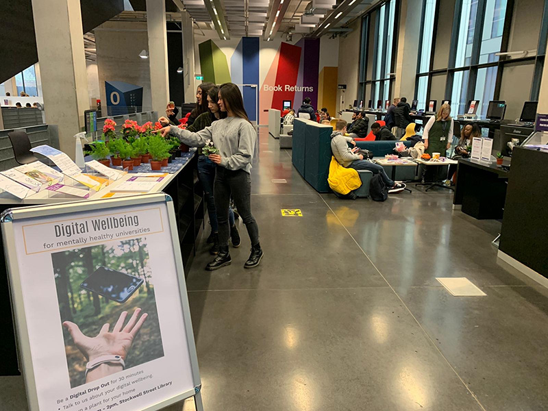 Our Digital Wellbeing library drop-in. Students stand at our display desk and explore the resources there. Beyond them students are using the library space to study and relax.