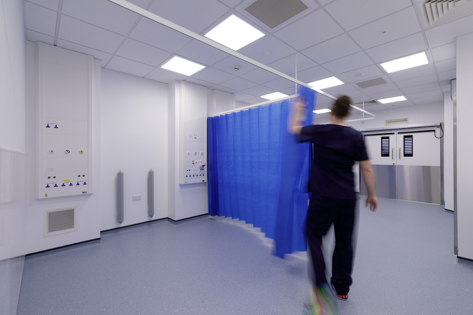 An example of how our CCU will look, the image shows an empty critical care facility with a person pulling a blue curtain across the space, in the distance are theatre doors