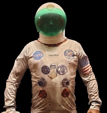 An old NASA astronaut suit, it is dirty white with lots of stickers on the chest. The helmet is white with a green visor. A dark face can be seen inside.