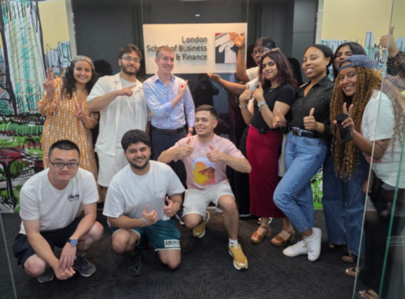A group of students and staff pose together in front of a sign for the London School of Business & Finance. They are standing and kneeling in a casual group formation, many making hand gestures such as thumbs‑up or pointing, and appear to be celebrating or marking an event. Colourful artwork decorates the walls around them.